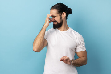 Bad smell. Portrait of man wearing white T-shirt standing pinching her nose with fingers to hold breath, disgusted by stinky intolerable smell. Indoor studio shot isolated on blue background.