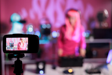 Fototapeta na wymiar Artist with pink hair standing at table in night club performing techno song using mixer console while filming music session with professional camera. Dj posting performance video on her channel