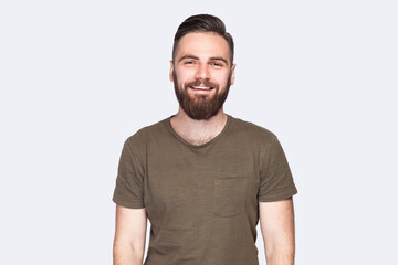 Fototapeta premium Portrait of handsome bearded man with stylish hairstyle wearing dark green T-shirt standing looking at camera with positive and optimism. Indoor studio shot isolated on light gray background.