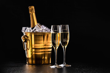 Two glasses of champagne and a bottle in a golden bucket with ice on a black background - obrazy, fototapety, plakaty
