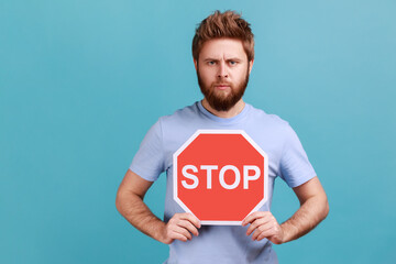 Portrait of handsome serious young adult bearded man holding octagonal Stop symbol, showing red...