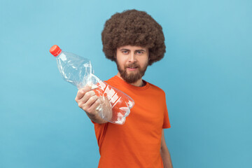 Portrait of responsible man with Afro hairstyle wearing orange T-shirt standing looking at camera with strict expression and showing plastic bottle. Indoor studio shot isolated on blue background.