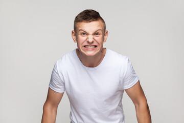 Obraz premium Portrait of handsome young teenager boy wearing T-shirt expressing aggressive emotions, screaming with hate and anger, frowning face. Indoor studio shot isolated on gray background.