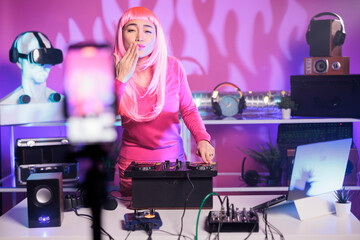 Artist with pink hair playing techno song at professional turntables while recording music process with smartphone camera. Dj person doing performance at nightclub with audio equipment