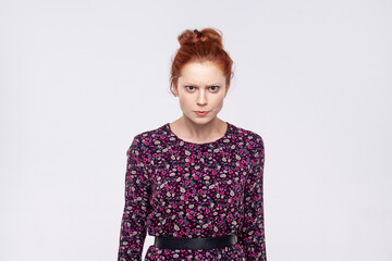 Portrait of angry unhappy young adult redhead woman wearing dress standing looking at camera, expressing aggression and hate. Indoor studio shot isolated on gray background.