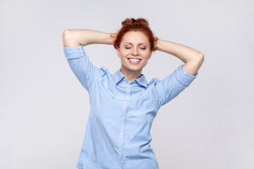 Portrait of beautiful attractive satisfied ginger woman wearing blue shirt standing with raised arms and closed eyes, smiling toothily. Indoor studio shot isolated on gray background.