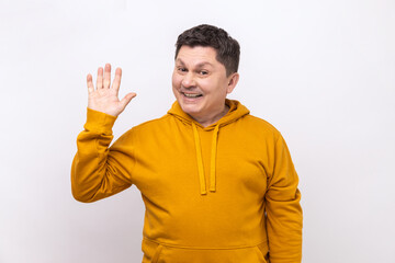 Friendly positive man greeting you rising hand and waving, saying hi, glad to see you, looking at camera with toothy smile, wearing urban style hoodie. Indoor studio shot isolated on white background.