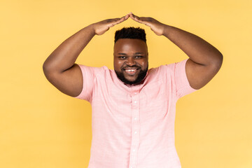 Portrait of attractive bearded man wearing pink shirt making house roof gesture and smiling,...
