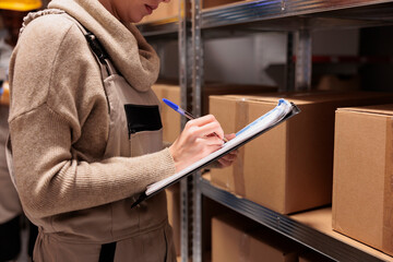 Delivery service worker marking received parcels in warehouse. Shipping company young woman employee working in storehouse, holding clipboard and writing in checklist close up