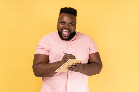 Portrait of man wearing pink shirt writing down in paper notebook, making to do list, having good mood, looking at camera with toothy smile. Indoor studio shot isolated on yellow background.