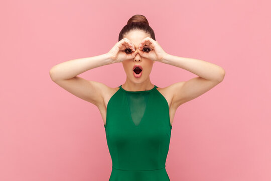 Portrait of funny shocked surprised woman standing with binoculars gesture, looking with open mouth, sees something astonished, wearing green dress. Indoor studio shot isolated on pink background.