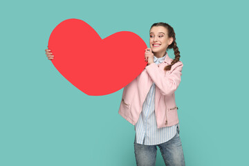 Fototapeta na wymiar Portrait of extremely happy cheerful teenager girl with braids wearing pink jacket, showing big red heart, looking away with toothy smile. Indoor studio shot isolated on green background.