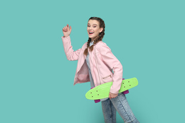 Portrait of extremely happy funny teenager girl with braids wearing pink jacket standing with raised arm, holding skateboard in hands. Indoor studio shot isolated on green background.