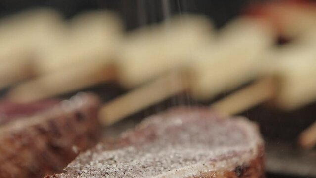 Medallions of sirloin steak being salted and seasoned on the grill over flames with coalho cheese in the background.