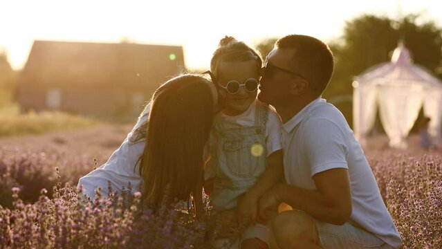 Family day. Happy young mom and dad holding hands and kissing their little daughter, enjoy relaxing in lavender field at sunset. Parents with kid enjoying summer holiday vacation. Children day.
