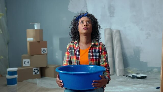Upset African American woman is holding blue bowl into which drops of water are dripping from ceiling leak. Female is calling on a mobile phone to master repairman or plumber and asking for help in