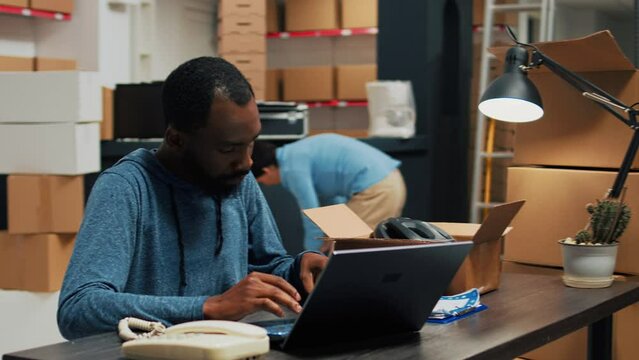 African american worker planning logistics to ship products, preparing order shipment with stock inventory information. Male owner working on laptop with packages from warehouse racks.