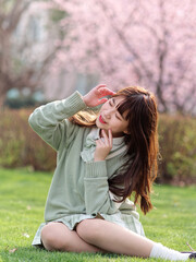 Obraz premium Outdoor portrait of beautiful young Chinese girl in Japanese JK style dress sitting with blossom cherry tree brunch background in spring garden, beauty, summer, emotion, expression and people concept.