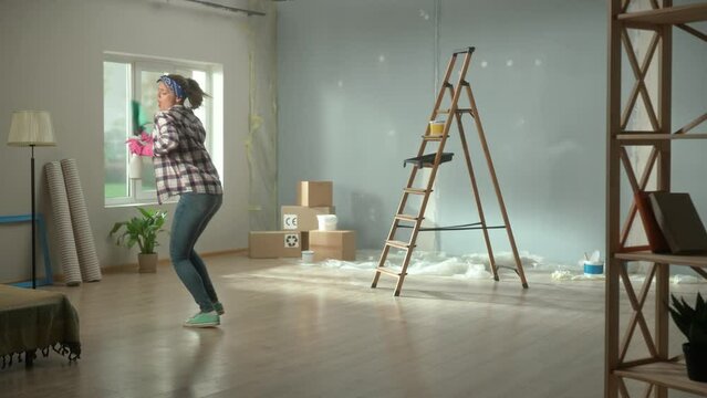 Young woman with rag and spray bottle singing and dancing merrily. Man joyfully singing into paint roller as if into microphone. Couple are having fun cleaning up an apartment during renovations.