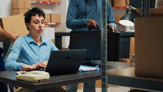 Diverse partners using laptop to do delivery logistics work in warehouse, preparing products order from storage room racks. Young people working on startup retail development, supply chain.
