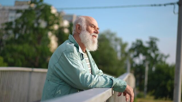 Side view thoughtful confident senior man looking around standing on bridge in urban city thinking. Confident handsome old Caucasian retiree enjoying warm sunny day in town outdoors