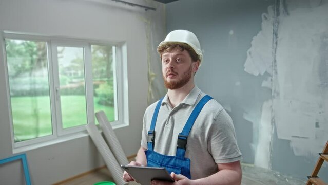 Male foreman is planning renovation project in an apartment using tablet. Redhead master man thinking, viewing and visualizing ideas against background of apartment in process of renovation.