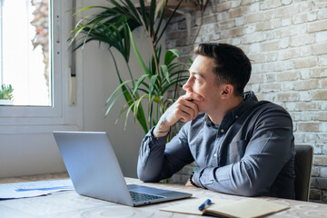 Thoughtful businessman touching chin, pondering ideas or strategy, sitting at wooden work desk with laptop, freelancer working on online project, student preparing for exam at home
