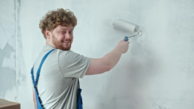 Foreman or painter in blue construction overalls is painting wall with white paint using paint roller. Redhead man is making repairs winking and showing thumbs up. Concept of repair, finishing works.