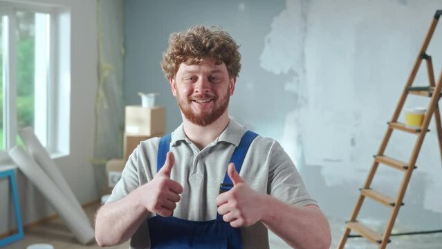 Repairman worker in blue overalls is smiling and showing thumbs up. Portrait of redhead man is showing gesture of recommendation and posing against backdrop of apartment in process of renovation.
