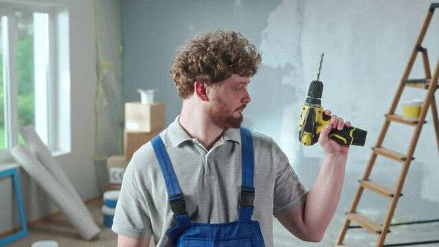 Repairman worker in blue construction overalls is demonstrating how an electric drill works. Portrait of redhead man is posing against backdrop of renovation apartment, ladder, cardboard boxes, window