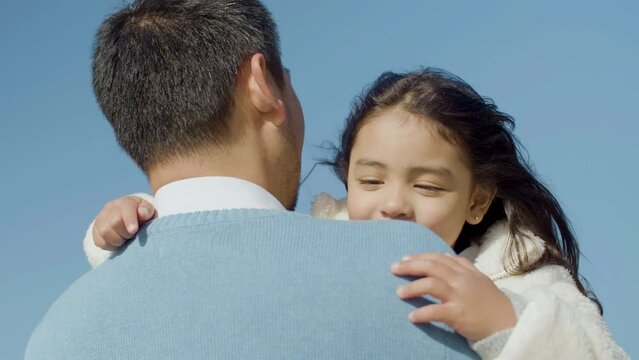 Happy Asian child spending time with her dad outdoors. Father holding little daughter in his arms and kissing her on cheek, then kid laughing. Family, parenthood, childhood concept