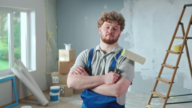 Repairman in blue overalls is looking confidently at camera and crossing arms with paint roller. Portrait of redhead man is posing against backdrop of renovation apartment, ladder, cardboard boxes