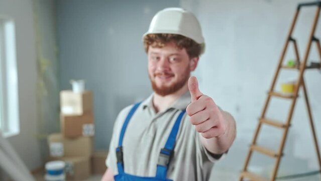 Repairman worker in blue overalls and white helmet is smiling and showing thumbs up. Portrait of redhead man is posing against backdrop of apartment in process of renovation.