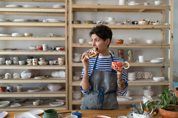 Relaxed woman in apron drink coffee from handmade ceramic cup and has snack in pottery studio....