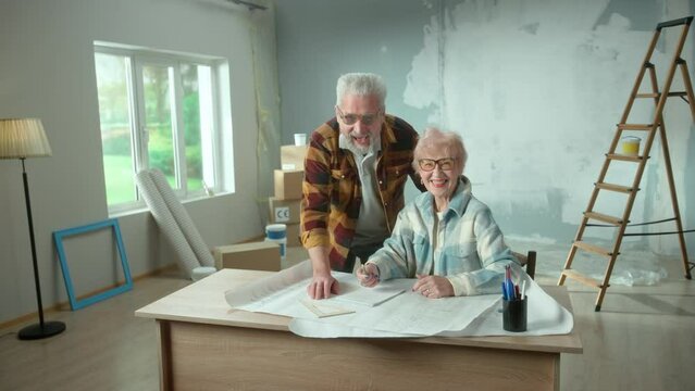 Elderly man and woman are looking through sheet with plan of an apartment and discussing renovation project. Happy aged couple is sitting at a table and planning the improvement of their home.