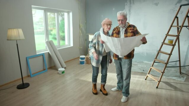 Elderly Man And Woman Are Looking Through Sheet With Plan Of An Apartment And Discussing Renovation Project. Aged Couple Is Planning Improvement Of Their Home. Concept Of Repair, Decoration, Interior