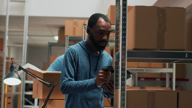 Small business owner working on distribution and inventory logistics, using scanner and tablet in warehouse space. Male worker scanning bar code on products boxes, selling goods. Handheld shot.