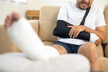 Man suffered pain from accident fracture broken bone injury with leg splints in cast neck splints collar arm splints sling support arm in living room. Social security and health insurance concept.