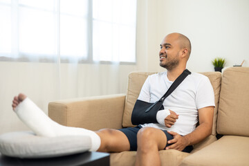 Happy man recovery from accident fracture broken bone injury with leg splints in cast neck splints collar arm splints sling support arm in living room. Social security and health insurance concept.