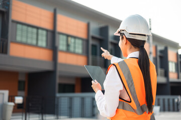 Confident engineer woman standing at modern home building construction. Architect with white safety helmet at construction site. Mechanic service factory Professional work job occupation in uniform.