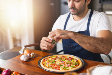 smiling young Caucasian man in apron and holding baking tray with fresh homemade pizza. Confident male chef holding cooked homemade pizza in kitchen. Italian delicious appetizer