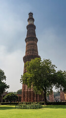 The world's unique tallest brick minaret in the Qutub Minar complex against the blue sky. Red tower with wavy walls and balconies. Lawn and trees in the foreground. India. Delhi