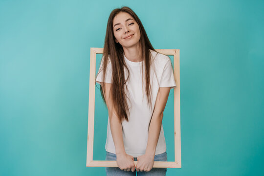 Cute cheerful young woman in white t-shirt and jeans holds frame for Canvas smiling wide stands over turquoise studio backdrop with empty space. Attractive Italian girl got into wooden frame. Mockup