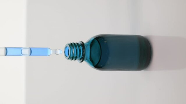 Abstract skin nourishing cosmetics formulating concept | Vertical extreme close-up crane shot of lab dropper drips blue oil into blue reagent bottle on grey background