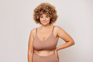Horizontal shot of busty body positive woman with curly hair looks confident and self loved poses...