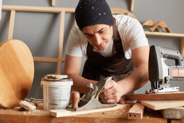 Indoor shot of concentrated man carpenter wearing apron and cap working with plane on wooden workplace background, making wooden furniture, enjoying his work in workshop, handmade wooden products.