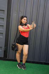 Fototapeta na wymiar Young attractive fashionable indian woman wearing casual short outfit and headphone taking selfie picture from smart phone holding handbag standing posing outdoor. Copy space.