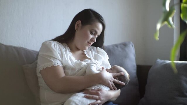 Madre sentada en el sof&aacute; amamantado a su beb&eacute; mientras lo acaricia para dormir realizando lactancia materna.