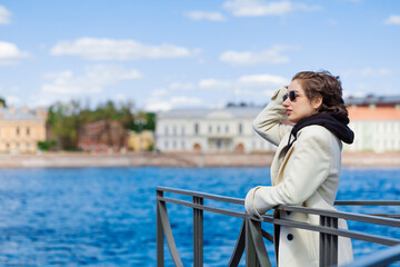 Beautiful young woman walks along embankment in white coat and black sweatshirt with sunglasses in sunny weather in spring. Tourism relaxes on the background of the river and the city