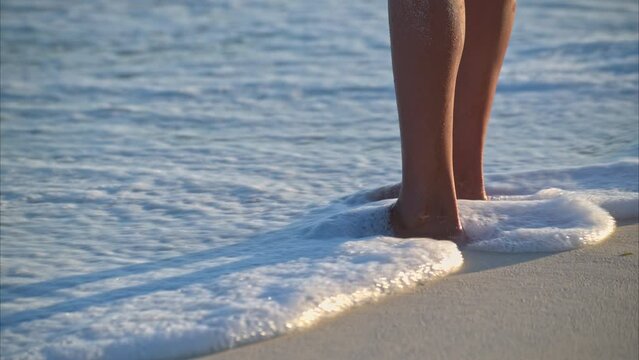 Slow motion of a pair of tanned thin woman legs standing on the sand having the waves splashing on her at the beach